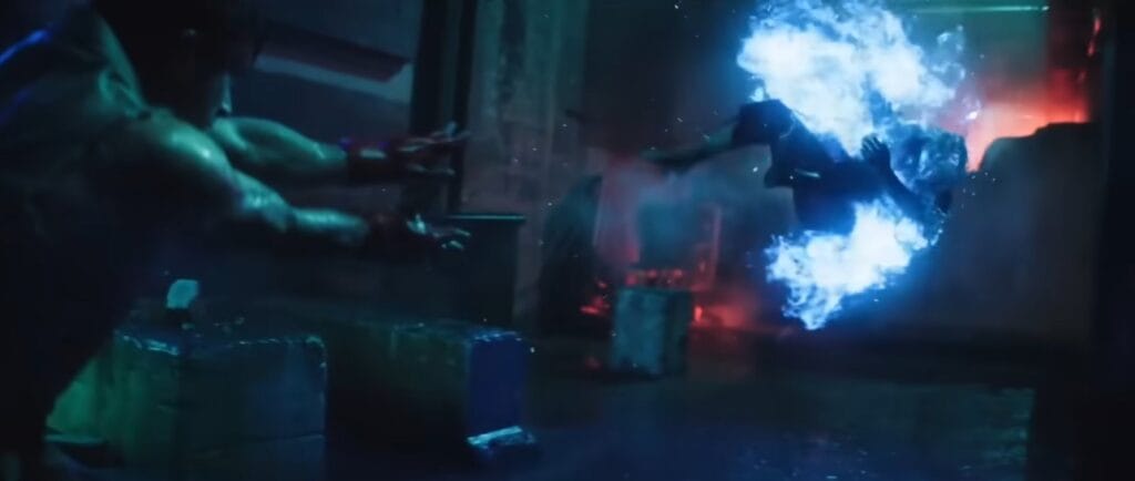 Ryu, vestindo kimono, lança um Hadouken de energia azul brilhante no Ken em um armazém escuro. Cena de luta do live-action de Street Fighter O Filme 2026 com iluminação dramática.