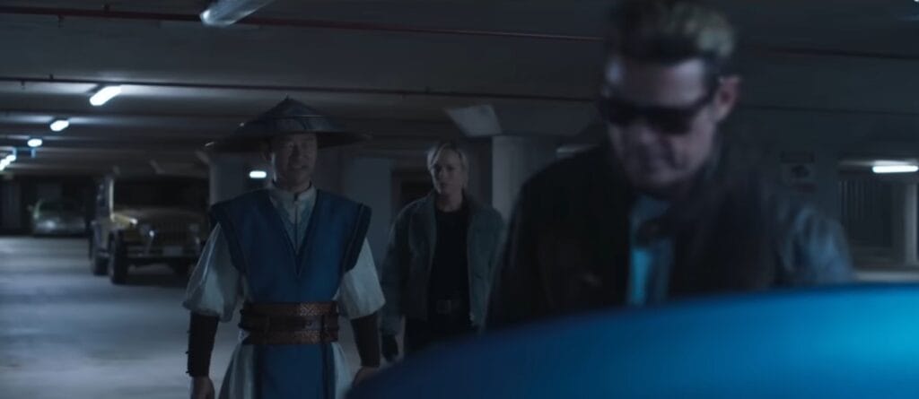 Raiden (Tadanobu Asano), vestindo seu traje azul e branco com o chapéu de palha clássico, caminha em um estacionamento subterrâneo ao lado de Sonya Blade (Jessica McNamee) em Mortal Kombat 2 (2026). Em primeiro plano e levemente desfocado, Johnny Cage (Karl Urban) aparece de óculos escuros, sinalizando o encontro dos defensores da Terra.