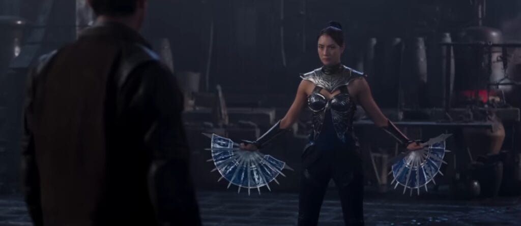 Kitana (Adeline Rudolph) em Mortal Kombat 2 (2026) em posição de combate segurando seus leques de guerra azuis e prateados com lâminas afiadas. Ela veste uma armadura de batalha azul detalhada e encara um oponente (Johnny Cage) em um ambiente sombrio.