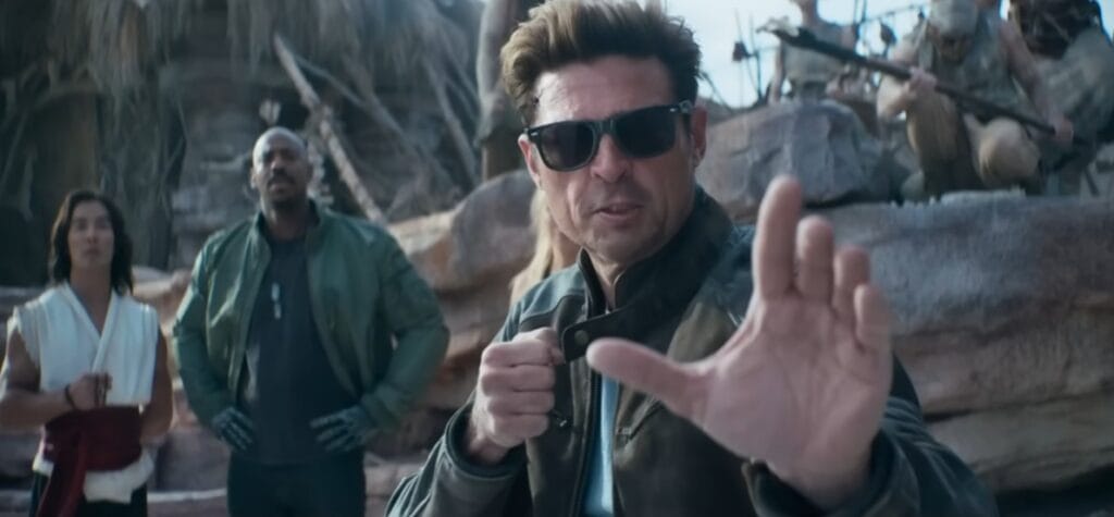 Johnny Cage (Karl Urban), de óculos de sol e pose de luta com a mão esquerda estendida, de pé no centro à frente de Liu Kang e Jax Briggs ao fundo, em um ambiente de ruínas de pedra no filme Mortal Kombat 2. SEO: Johnny Cage (Karl Urban), cena do filme Mortal Kombat 2, adaptação live-action de videogame, pose de luta.