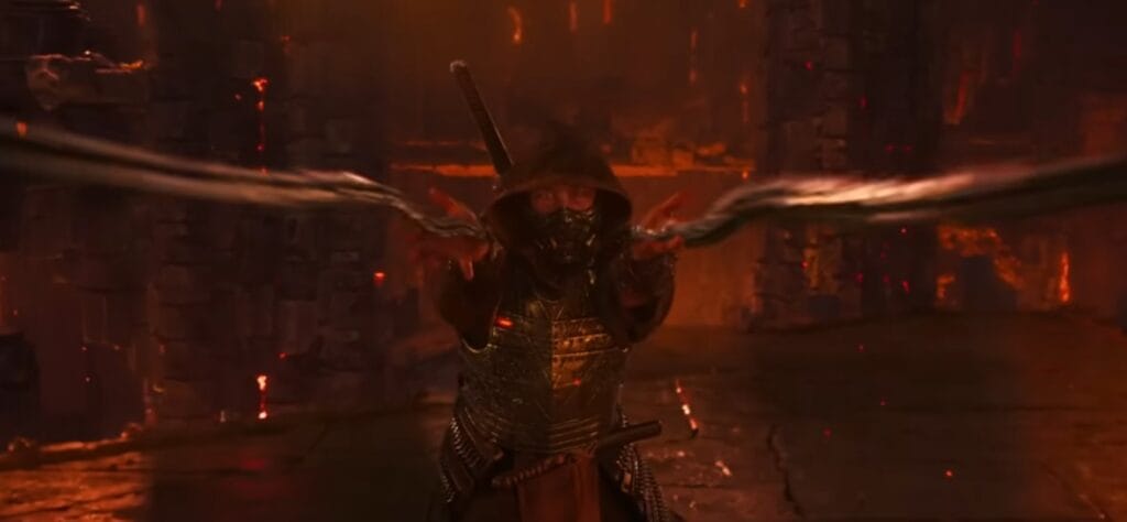 Scorpion (Hiroyuki Sanada) em Mortal Kombat 2 (2026) disparando suas icônicas correntes com kunai em direção à câmera. Ele veste sua armadura amarela dourada com detalhes em preto e máscara ninja, em um cenário infernal com lava e fumaça ao fundo.