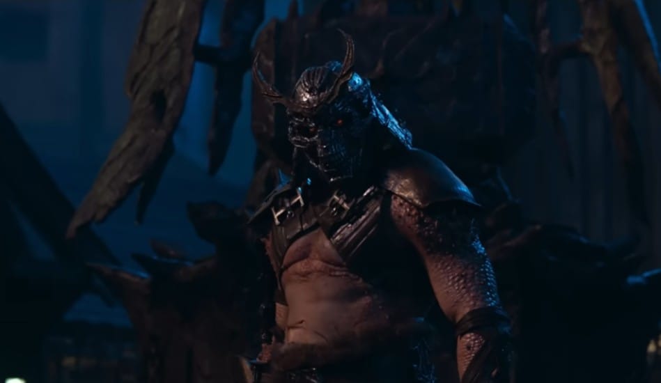 Shao Kahn (interpretado por Martyn Ford) em Mortal Kombat 2 (2026), vestindo seu icônico capacete de caveira com chifres e olhos vermelhos brilhantes. O vilão aparece em um ambiente sombrio com iluminação azul, destacando sua armadura de batalha e seu físico massivo e imponente.