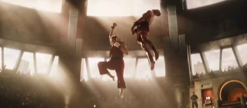 Ken, de kimono vermelho, executa um Shoryuken flamejante ascendente contra um oponente no ar em uma arena de torneio iluminada. Cena de luta do live-action Street Fighter O Filme 2026.