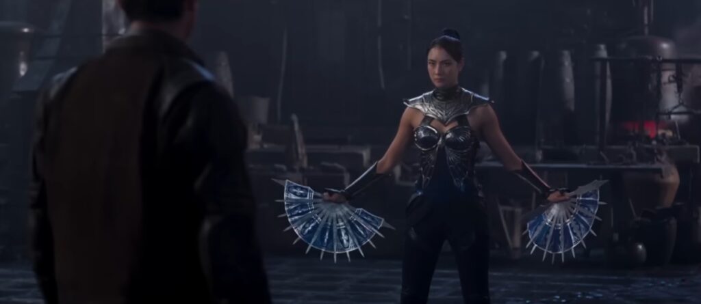 Kitana (Adeline Rudolph) em Mortal Kombat 2 (2026) em posição de combate segurando seus leques de guerra azuis e prateados com lâminas afiadas. Ela veste uma armadura de batalha azul detalhada e encara um oponente (Johnny Cage) em um ambiente sombrio.